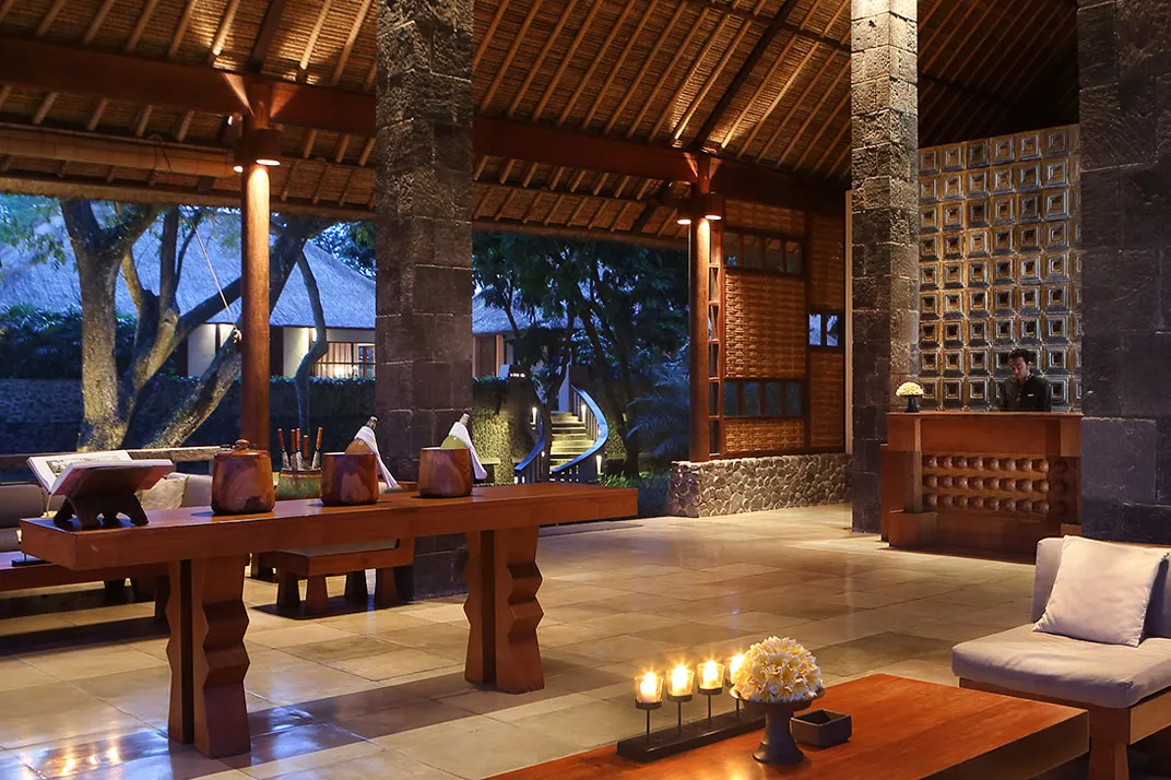 The open-air lobby at Alila Ubud at dusk, stone columns under a high alang-alang roof