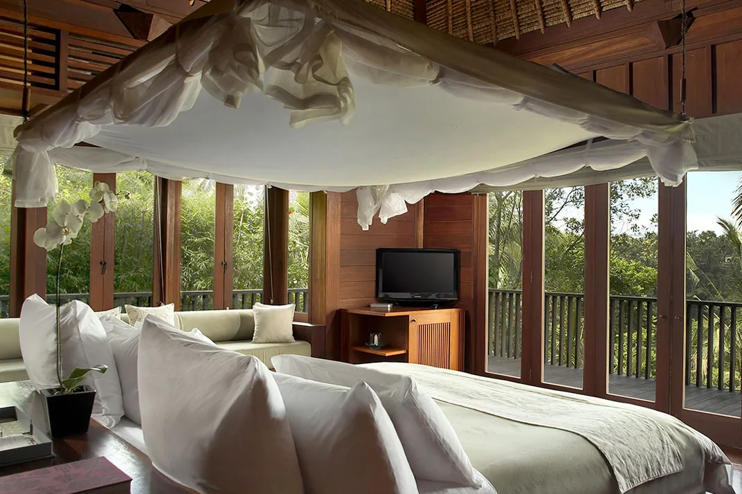A villa bedroom at Alila Ubud under a thatched ceiling, with glass doors onto a terrace above the jungle