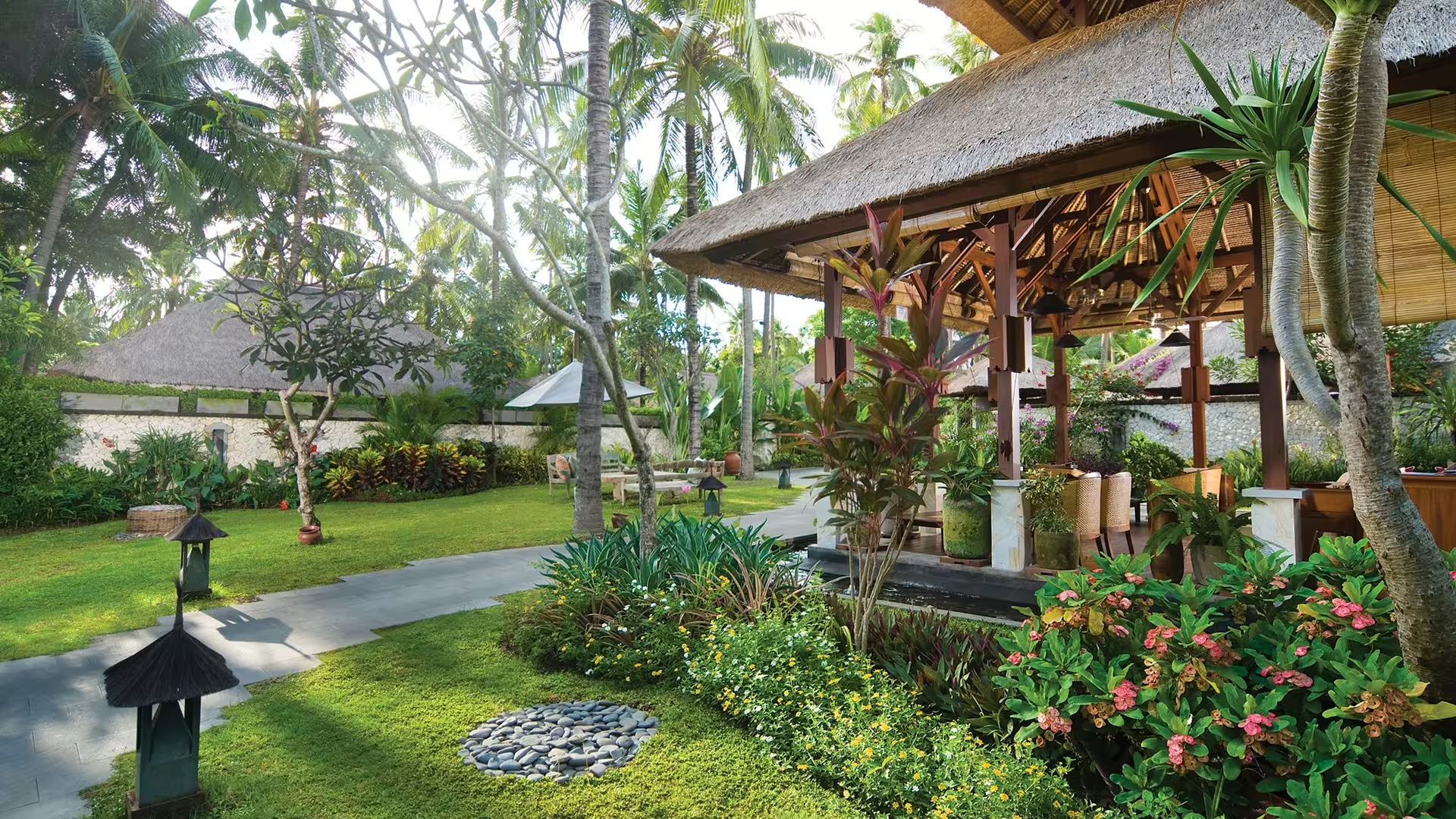 A stone path through Jimbaran Puri's gardens, leading to a thatched pavilion among frangipani and tropical plants