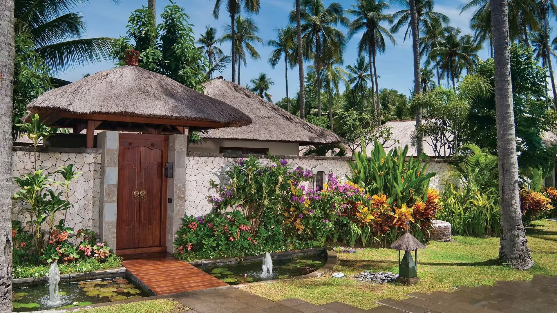 A garden entrance at Jimbaran Puri, with a koi pond, thatched roofs, and a traditional wooden gate