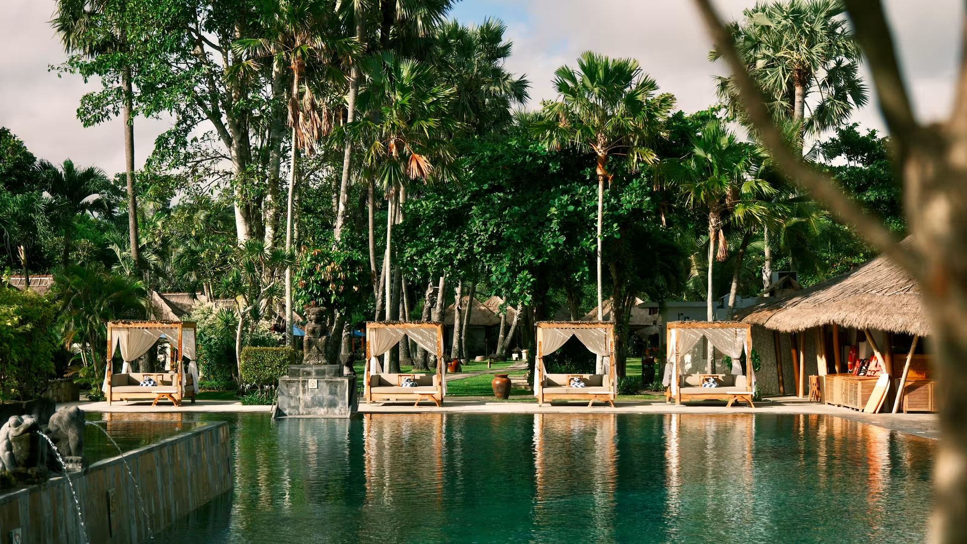 The main pool at Jimbaran Puri at golden hour, with thatched daybeds along the water's edge