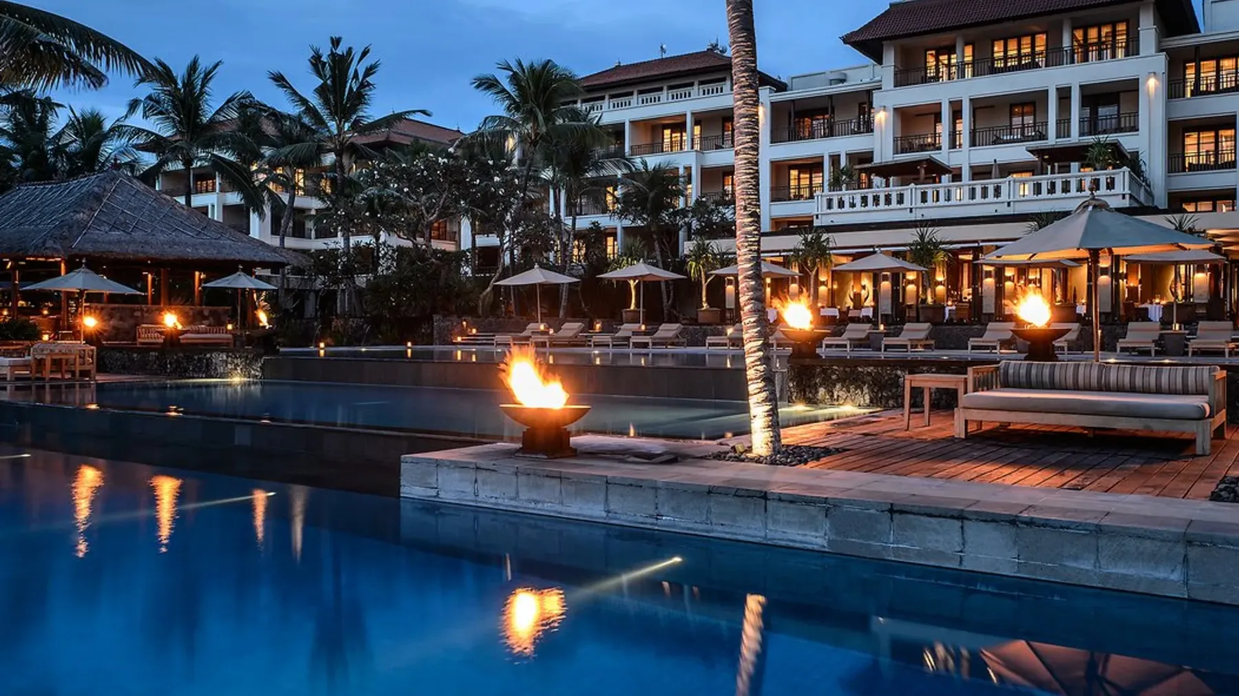 Fire bowls lit beside the infinity pools at The Legian at dusk, the hotel glowing behind