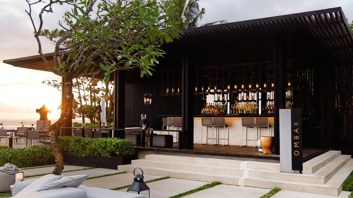 A cliffside table at Soori Bali at sunset, the temple-topped rock visible along the coastline