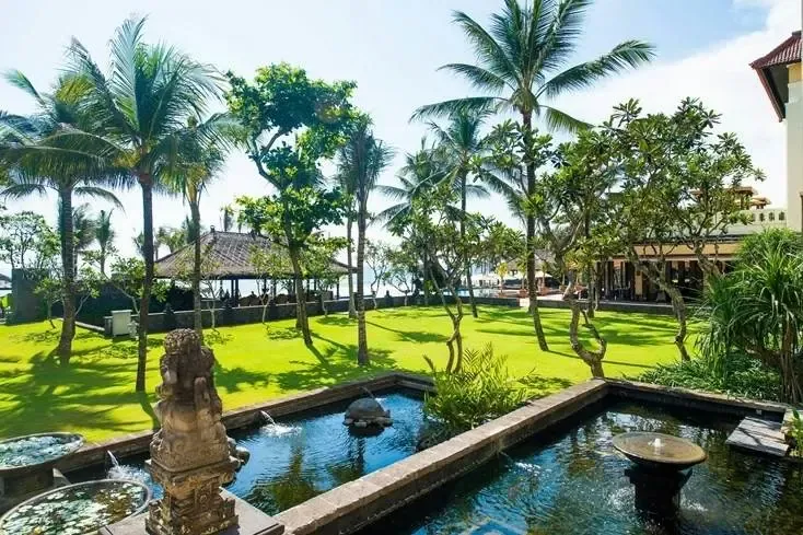 A koi pond and Balinese stone statue in the garden at The Legian, with the beach visible beyond the lawn