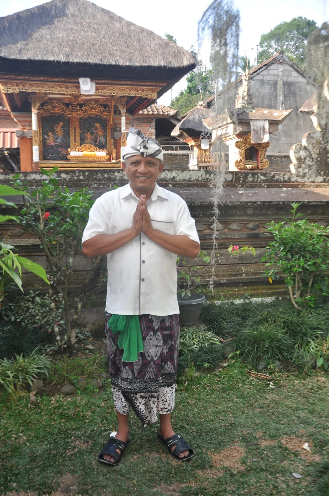 Wayan in traditional Balinese dress, hands pressed together in greeting, in front of a carved pavilion